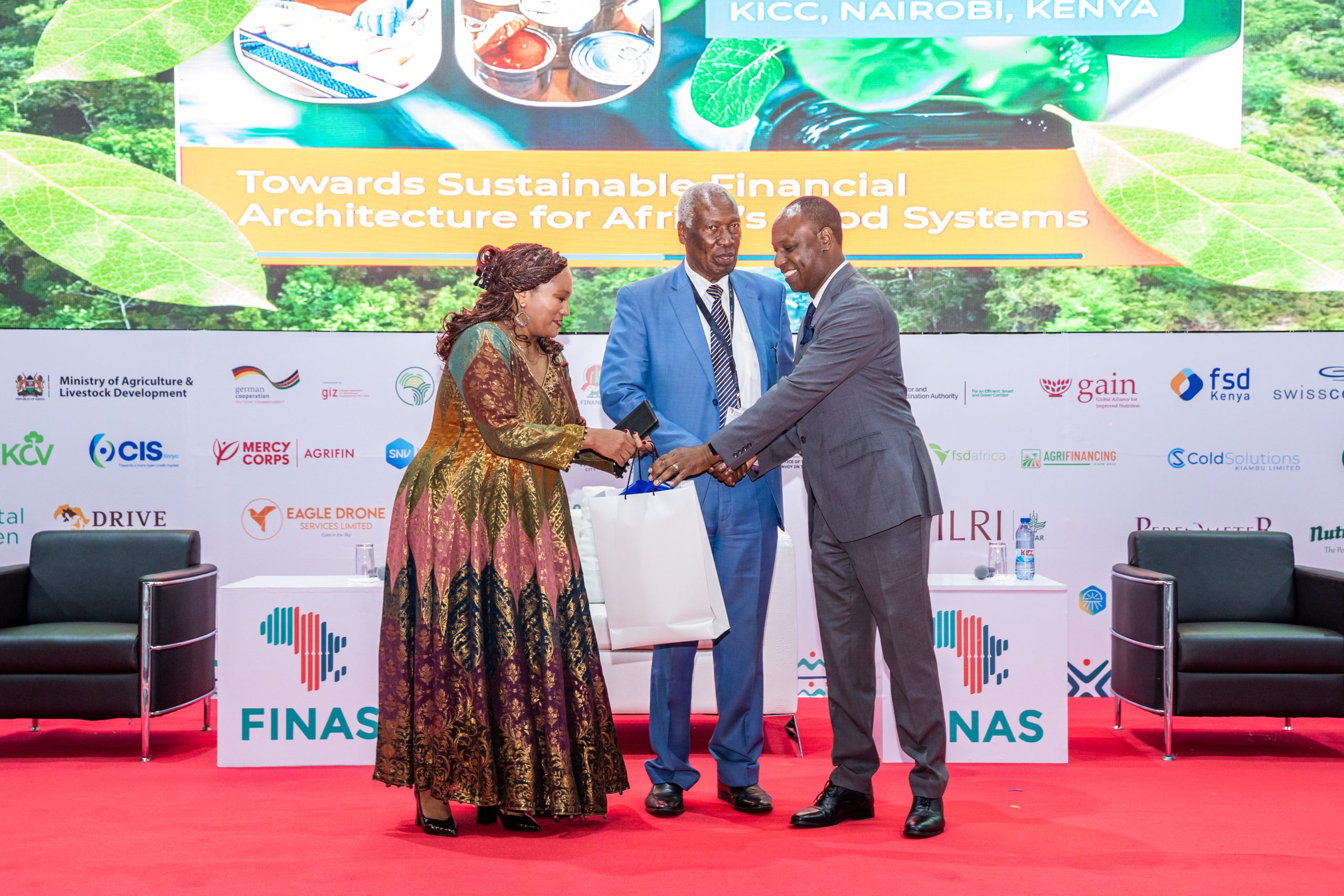 Dr. Charity Mutegi (l) and Tharaka University Chancellor, Prof. Ratemo Michieka (c) presenting a gift to Livestock Development PS, Jonathan Mueke (r) during the opening ceremony of FINAS